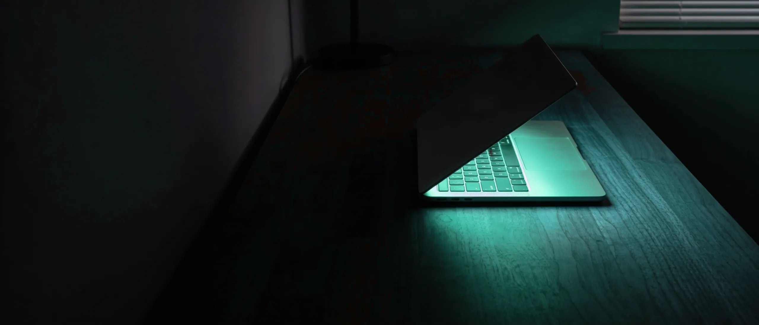 A laptop glowing teal on a desk in a dark room at night